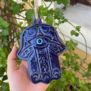 Può includere: Una mano di Hamsa in ceramica blu scuro con un ciondolo blu malocchio. La mano di Hamsa è un simbolo di protezione e buona fortuna.