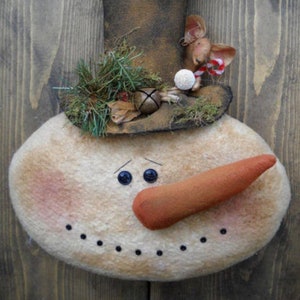 Pode incluir: Um enfeite de boneco de neve de feltro feito à mão com um chapéu-coco marrom, um nariz de cenoura e olhos de botão pretos. O boneco de neve é decorado com ramos verdes, musgo e um pequeno rato de feltro branco que usa um bastão de caramelo vermelho e branco.