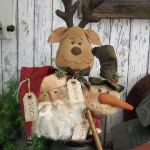 Pode incluir: Um trio de decorações de Natal feitas à mão com um Papai Noel, um boneco de neve e uma rena. As figuras são feitas de tecido e têm um estilo rústico e vintage. O Papai Noel usa um chapéu vermelho e tem uma barba branca. O boneco de neve usa um chapéu preto e tem um nariz de cenoura. A rena tem chifres marrons e uma cara marrom. As figuras estão todas em pé em um pote marrom.