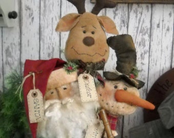 May include: A trio of handmade Christmas decorations featuring a Santa, a snowman, and a reindeer. The figures are made of fabric and have a rustic, vintage style. The Santa is wearing a red hat and has a white beard. The snowman is wearing a black hat and has a carrot nose. The reindeer has brown antlers and a brown face. The figures are all standing in a brown pot.