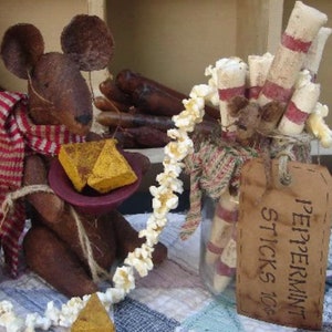 以下が含まれることがあります： 赤と白のストライプのストールを巻いた茶色のネズミの置物が、黄色のチーズの角が入ったボウルを持っています。白と赤のペパーミントスティックが入った瓶は、ポップコーンのガーランドと「Peppermint Sticks 10c」と書かれた茶色のタグで飾られています。