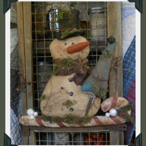 May include: A handmade primitive snowman with a black top hat, a red nose, and a blue scarf. The snowman is sitting on a wooden shelf with a candy cane and white beads. The snowman is decorated with moss and has the words "Merry Christmas" written on it.