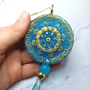 Puede incluir: Colgante redondo azul y dorado con diseño de rueda. El colgante tiene un borde dorado y una rueda dorada en el centro. Una cuenta azul, una cuenta dorada y una borla azul y dorada cuelgan del colgante.