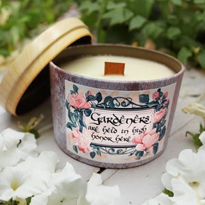 May include: A purple and white candle jar with a wooden lid and a floral design. The label reads "Gardeners are held in high honor here".