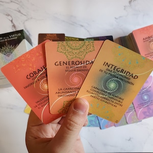 May include: Close-up of three orange, yellow and pink oracle cards with Spanish text. The cards feature intricate mandala designs and are part of a larger deck. The text on the cards reads "Coraje", "Generosidad", and "Integridad".