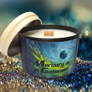 May include: A black candle with a wooden wick and a light-colored wooden lid. The candle's label features a swirling blue and green design with the text "Mercury is in Gatorade."