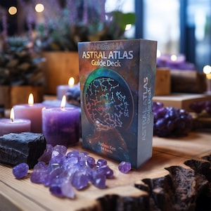 An oracle deck box with the words 'PANDEMONIUM: ASTRAL ATLAS Guide Deck' on the front. The box is sitting on a wooden surface with purple crystals and candles. The box has a design of a galaxy with stars and constellations.