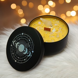 May include: A black candle jar with a gold lid and a yellow candle with white and gold glitter inside. The candle jar has a design of a sun and moons on the lid.