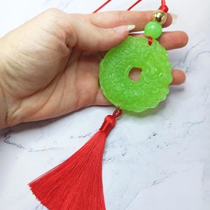 May include: A jade green pendant with a central hole, intricate carvings, and a red tassel. The pendant is suspended from a red cord, with a green bead and a gold-coloured bead. The design is likely inspired by Asian cultural motifs.