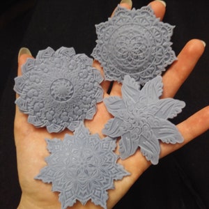 Puede incluir: Cuatro moldes de silicona grises decorativos en forma de mandala. Los moldes tienen forma de flor, estrella y dos círculos.