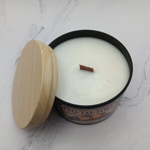 May include: A black candle jar with a wooden lid and a wooden wick. The candle is white and has the text "PO-TAY-TOES" printed on the jar.