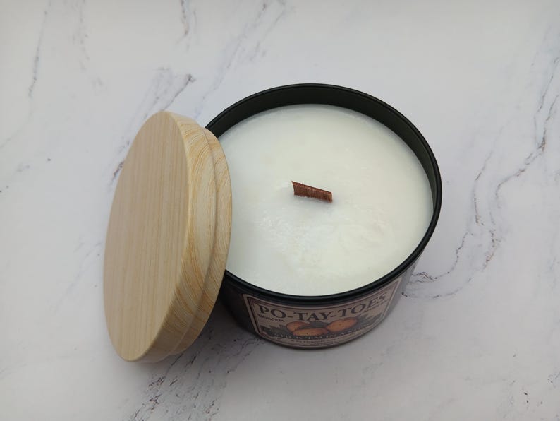 May include: A black candle jar with a wooden lid and a wooden wick. The candle is white and has the text "PO-TAY-TOES" printed on the jar.