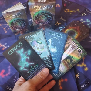 A set of astrology oracle cards. The cards feature illustrations of constellations, animals, and other celestial imagery. The cards are titled 'CERVUS', 'URANUS', 'WATER', and 'BEAUTY'. The cards are held in a fan shape over a blue astrology cloth.