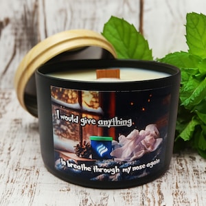 May include: Black candle with a wooden lid and a label that reads "I would give anything to breathe through my nose again". The label also features a winter scene with a window, snow, and a tissue box. The candle is sitting on a wooden surface.