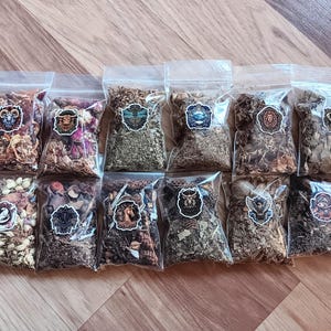 Puede incluir: Una colección de pequeñas bolsas de plástico transparente llenas de hierbas secas y productos botánicos. Cada bolsa tiene una pegatina con un símbolo del zodiaco. Las bolsas están dispuestas sobre una superficie de madera, mostrando una variedad de colores y texturas.