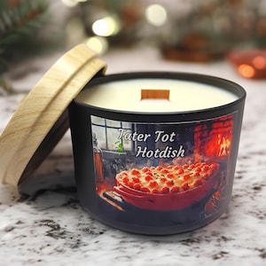 Puede incluir: Un tarro de vela negro con una tapa de madera y un interior de cera blanca. El tarro tiene una etiqueta con una imagen de una fuente para hornear llena de croquetas de patata y el texto "Tater Tot Hotdish".