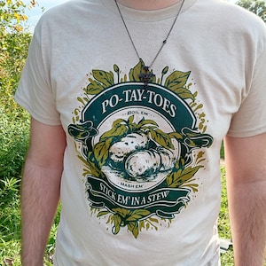 Puede incluir: Camiseta beige con un diseño verde y blanco. El diseño presenta el texto "PO-TAY-TOES" y "Stick 'em in a stew", con ilustraciones de patatas y follaje. Se lleva un collar negro.