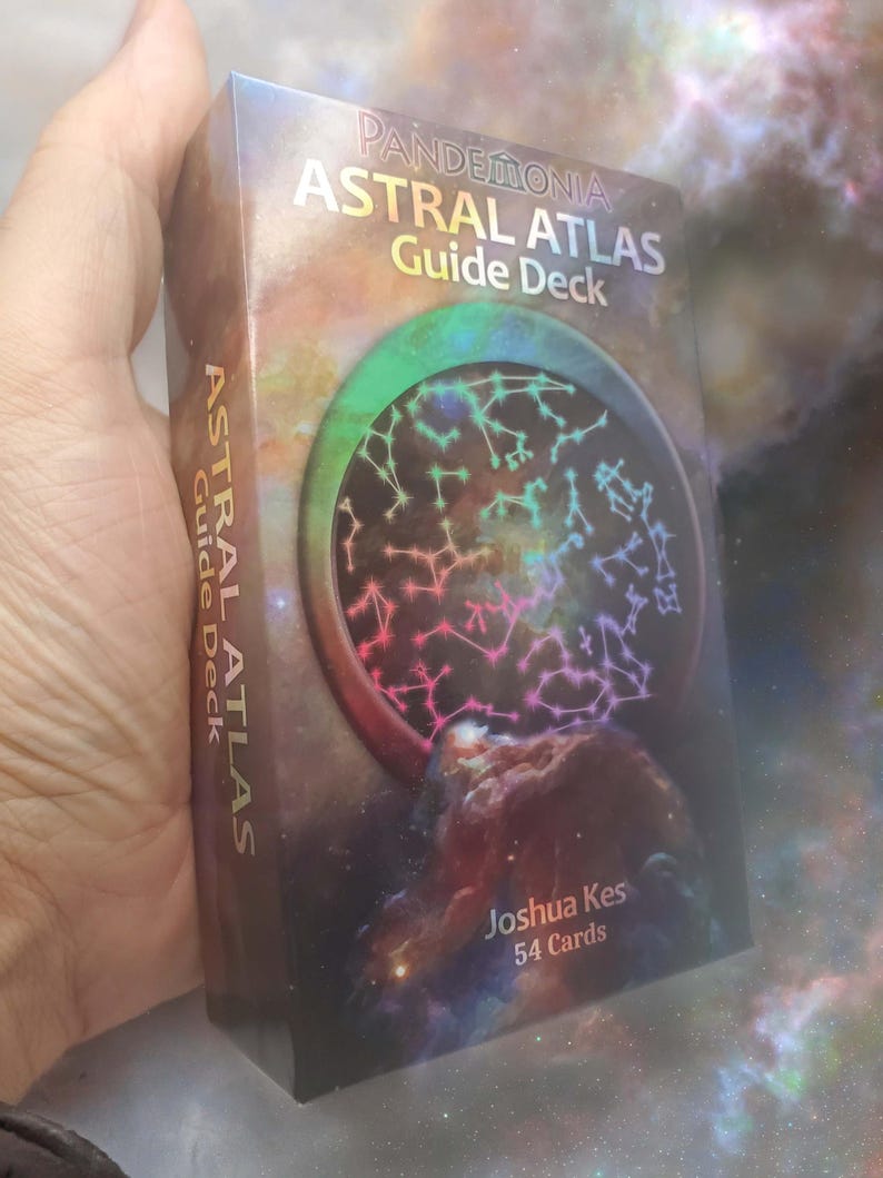 May include: A rectangular deck of cards box titled Pandemonia Astral Atlas Guide Deck by Joshua Kes. The box features a colorful nebula background with a circular graphic containing white constellation patterns and the text 54 Cards on the front.