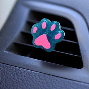 May include: A teal and pink paw print car air vent clip. The paw print is a translucent teal colour with pink pads. The clip is designed to attach to a car's air vent.