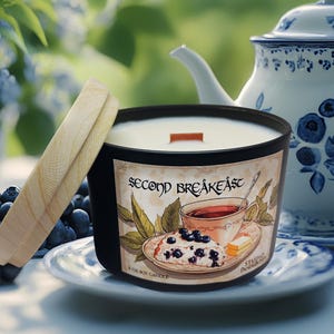 May include: A black candle with a wooden lid and a label that reads "Second Breakfast". The label features a drawing of a teacup, blueberries, and a slice of cake. A blue and white teapot is in the background.