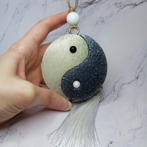 May include: Yin Yang shaped ornament with a white and dark blue speckled design. The ornament has a white tassel and a white bead at the top, attached to a natural fiber cord. The ornament is held by a hand.