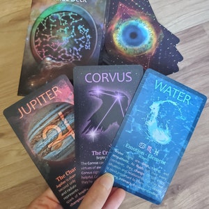 Three astrology oracle cards from the Astral Atlas Guide Deck held for display in front of the deck box and a pile of other cards on a wooden surface. The cards are Jupiter, Corvus, and Water. The cards are orange, purple, and blue.