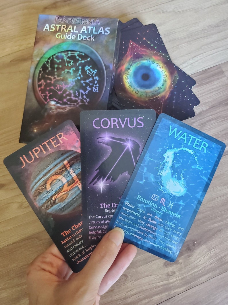 Three astrology oracle cards from the Astral Atlas Guide Deck held for display in front of the deck box and a pile of other cards on a wooden surface. The cards are Jupiter, Corvus, and Water. The cards are orange, purple, and blue.