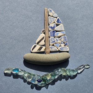 Könnte beinhalten: Ein handgefertigtes Segelboot aus Meeresglas und Keramikscherben. Das Segel besteht aus blau-weiß gemusterten Keramikteilen mit einem Holzmast. Der Rumpf ist ein glatter, grauer Stein, und die Wellen werden durch klares und blaues Meeresglas dargestellt.