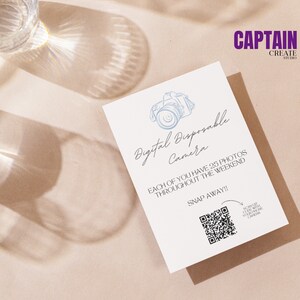 May include: A white card with blue text and a blue line drawing of a camera. The text reads "Digital Disposable Camera" and "Each of you have 25 photos throughout the weekend. Snap Away!" A QR code is in the bottom right corner with the text "Scan QR code with your phone camera". The card is on a brown surface.