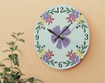 May include: A light blue round clock with black hands and a floral design. The clock face has a purple flower in the center and a wreath of smaller flowers around the edge. The numbers 12, 3, 6, and 9 are printed on the clock face.