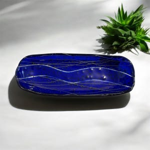 May include: A rectangular, deep blue glass dish with a wavy white design. The dish has a glossy finish and a dark border. A small green plant is visible in the background, adding a natural element to the composition.