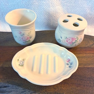 May include: A three-piece bathroom accessory set in cream-colored ceramic, featuring a tumbler, a four-hole toothbrush holder, and an oval soap dish. Each piece is decorated with a delicate floral pattern of pink roses and green leaves.