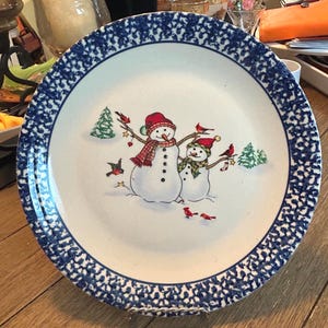 May include: A round ceramic dinner plate featuring a white center with a festive illustration of two snowmen wearing scarves and hats, surrounded by small green pine trees and red birds. The plate has a wide, dark blue speckled border rim.
