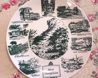 May include: A white ceramic souvenir plate featuring dark green illustrations of New Hampshire landmarks including the Old Man of the Mountains, State House, Dartmouth Hall, and various historic sites with their names printed in black text.
