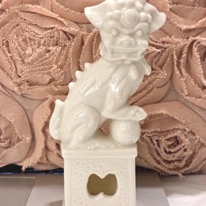 May include: A white ceramic Foo Dog figurine standing on a rectangular pedestal base. The mythical creature has a detailed mane, a fierce expression, and one paw resting on a spherical ball. The base features a central cutout and textured sides.