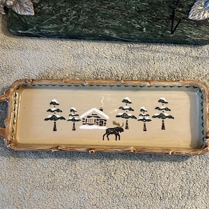 May include: Rectangular ceramic serving tray with a winter scene. The beige tray features a cabin, trees, and a moose illustration. The rim and handles have a wood-like design.