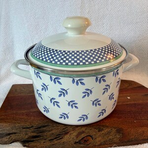 May include: A white enamel pot with two side handles and a matching lid featuring a cream-colored knob. The pot is decorated with a repeating pattern of small blue leaves, while the lid displays a blue and white checkered border pattern.