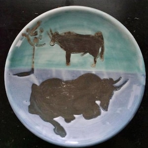 May include: A ceramic plate with a blue and white glaze. The plate features a brown silhouette of a bull standing on a light blue background and a brown silhouette of a bull lying down on a white background.