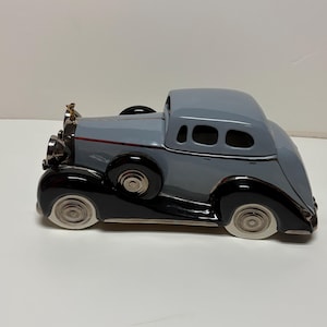 May include: A ceramic vintage car figurine in shades of gray and black. The car has a classic design with a rounded body, detailed headlights, and white-walled tires. A gold-colored ornament sits on the hood.