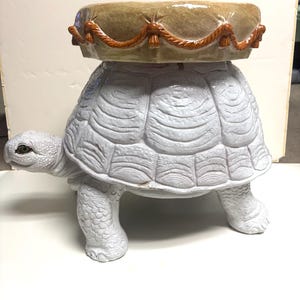 May include: A white ceramic turtle-shaped garden stool. The turtle has a detailed shell and legs, with a round, tan-colored top. The top is trimmed with a brown rope design. A unique decorative piece for outdoor or indoor use.