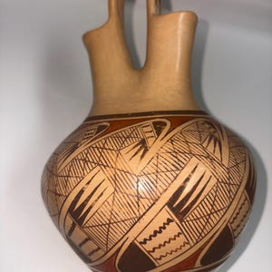 May include: A tan ceramic vessel featuring a double-spout design with a central arched handle. The rounded body is decorated with intricate geometric patterns in dark brown and reddish-brown, including cross-hatching and zigzag lines.