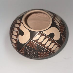 May include: A round, low-profile ceramic pot with a wide opening. The exterior features a matte finish with a geometric pattern in black, terracotta, and cream. The design includes repeating leaf-like shapes, solid black curves, and intricate cross-hatched sections.