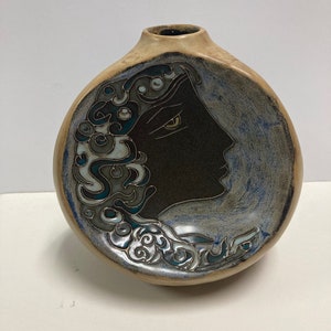 Może przedstawiać: Ceramiczny wazon o okrągłym kształcie z ozdobnym wzorem twarzy. Twarz jest pomalowana na czarno z złotym okiem i otoczona niebieskimi i białymi wirami.