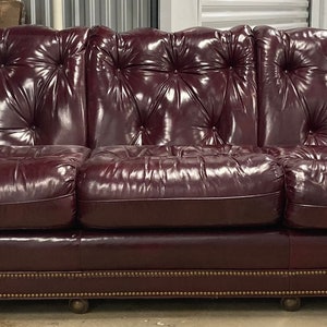 Op de afbeelding: Een bordeauxrode leren bank met gestoffeerde bekleding en een studded rand. De bank heeft een klassiek ontwerp en is perfect voor een woonkamer of studeerkamer.