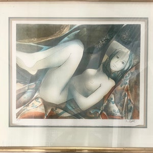 May include: Framed print of a nude figure reclining on patterned fabric. The artwork features soft colors, with the figure rendered in pale tones against a backdrop of muted blues, browns, and oranges. The print is in a gold-colored frame.