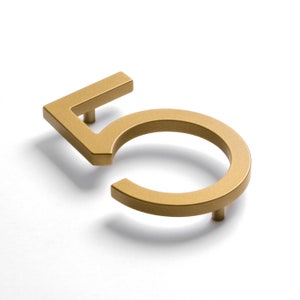 May include: A three-dimensional house number five finished in a matte gold tone. The numeral features a clean, geometric sans-serif font with sharp angles and a circular base. Two small cylindrical mounting posts are visible on the underside.