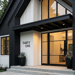 May include: A modern house exterior featuring black vertical siding and white board-and-batten accents. The entrance has a large glass door with black frames, a black metal awning, and the text THIRTY ONE mounted on the white wall near a black planter.