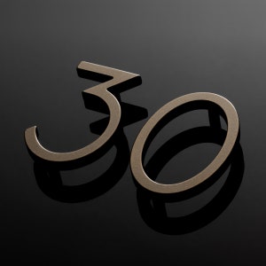 Pode incluir: Uma placa de número de casa de metal de cor bronze com o número "30" em uma fonte moderna e estilizada.
