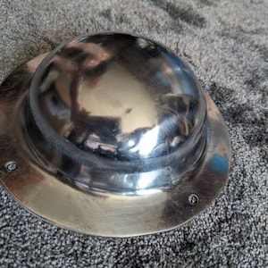 May include: A silver metal shield with a domed centre. The shield has a smooth, reflective surface.