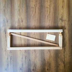 May include: A wooden frame with two dowels and a small piece of paper inside. The frame is made of light wood and has a simple design.